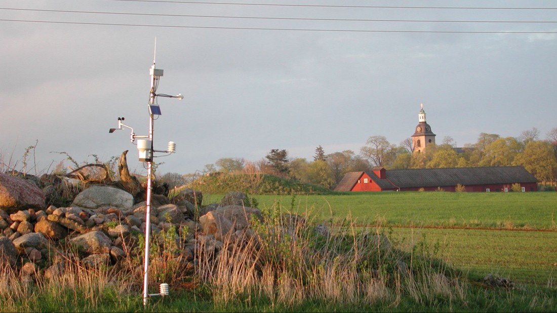 Väderstation vid en åkerholme. I bakgrunden syns en röd lada och ett kyrktorn.